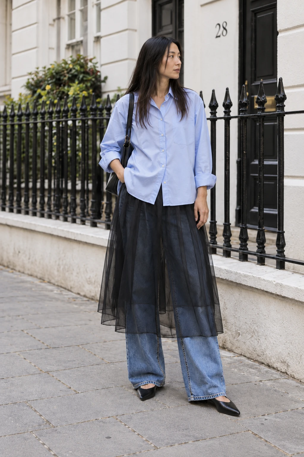 Keep the jeans. The jeans stay on under the black tulle skirt, and both hems remain visible. The loose blue shirt gives all that layering some breathing room. A skirt over trousers is easier to take seriously when the trousers are this ordinary.
